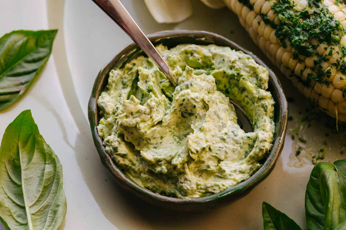 Bowl of basil butter with a spoon next to an ear of grilled corn brushed with melted basil butter.
