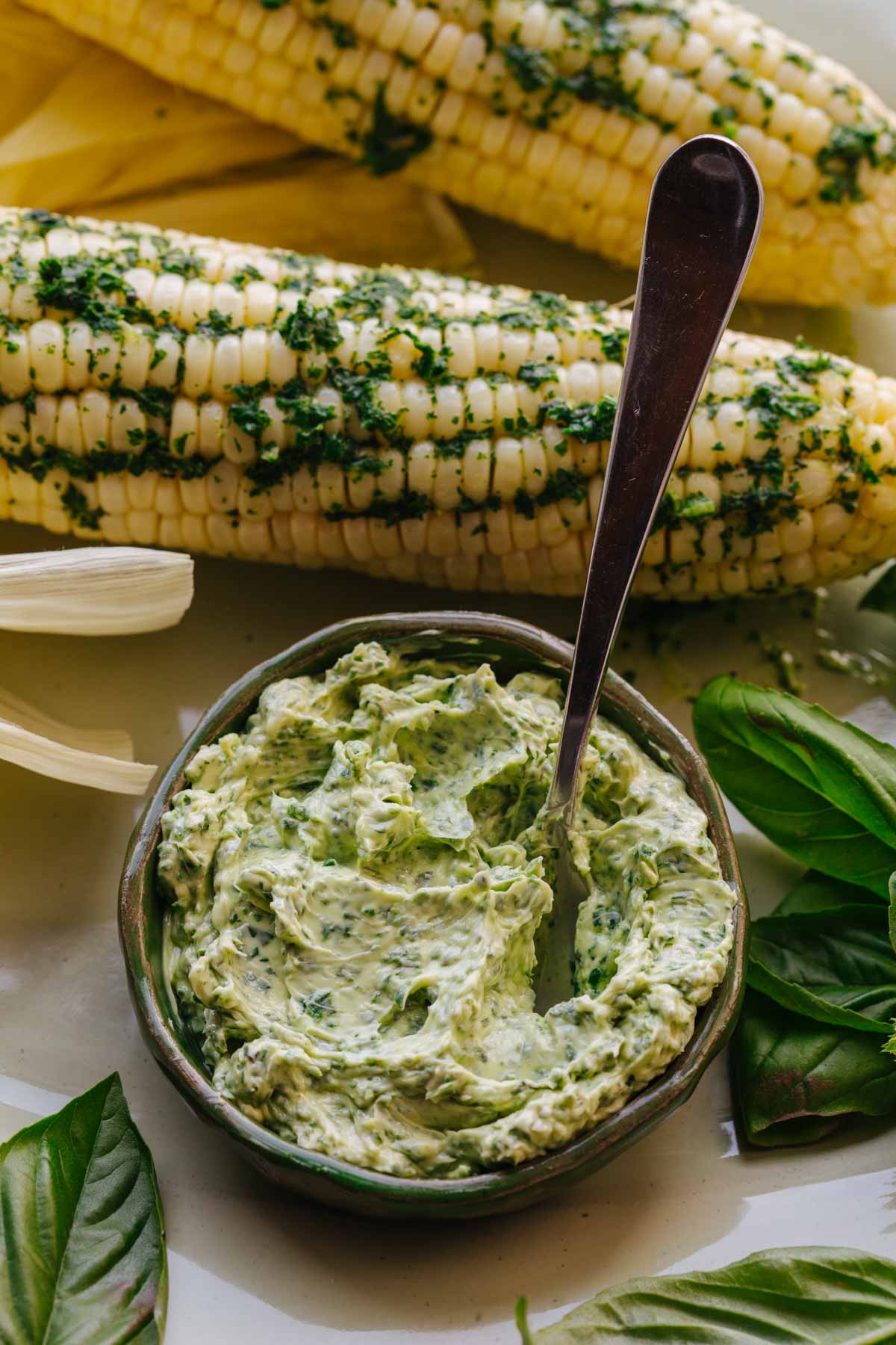 Two ears of corn topped with melted basil butter, with a bowl of basil butter and a spoon in the foreground.