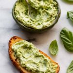 Slice of toasted bread spread thickly with basil butter, next to a bowl of basil butter and fresh basil leaves.