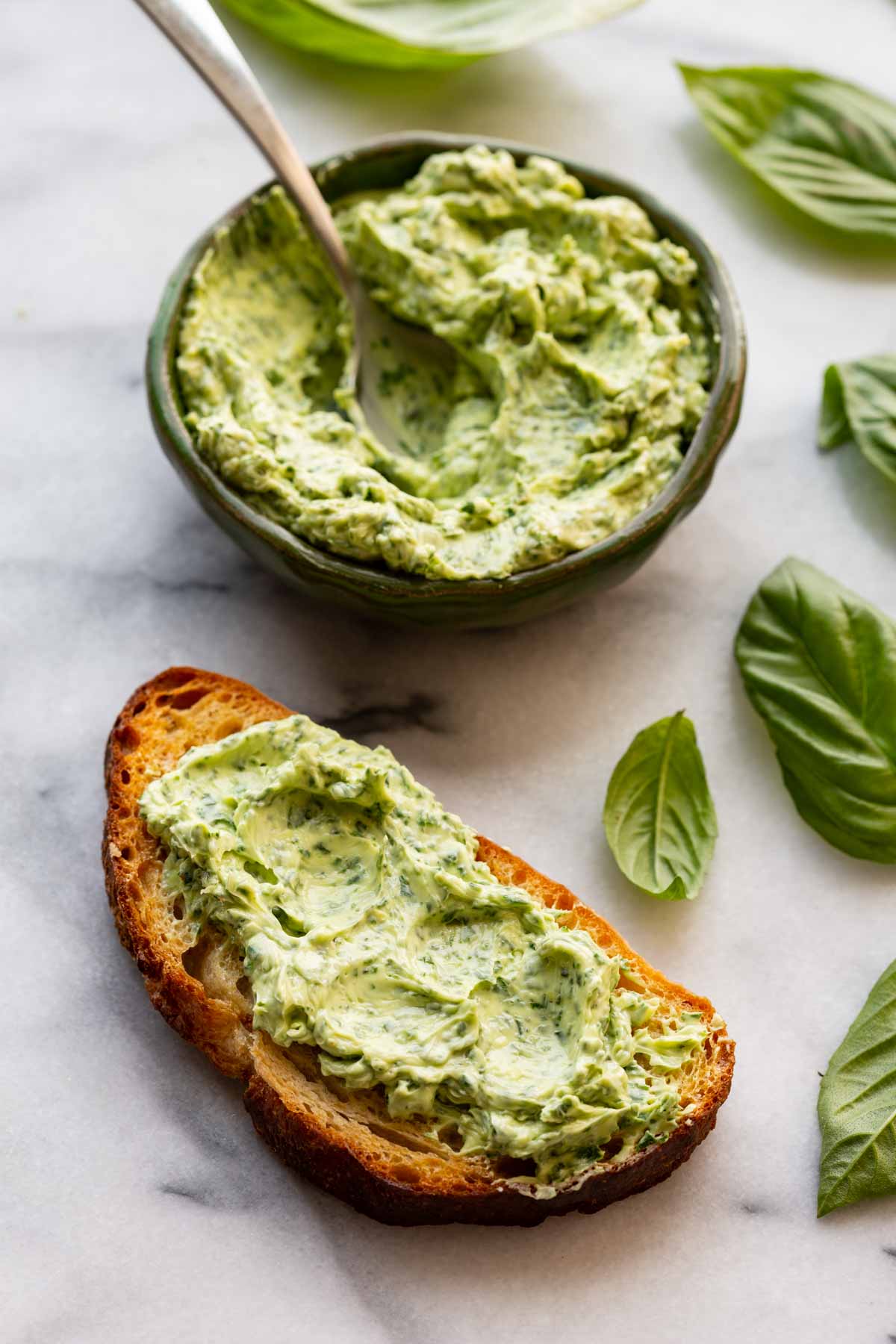 Slice of toasted bread spread thickly with basil butter, next to a bowl of basil butter and fresh basil leaves.