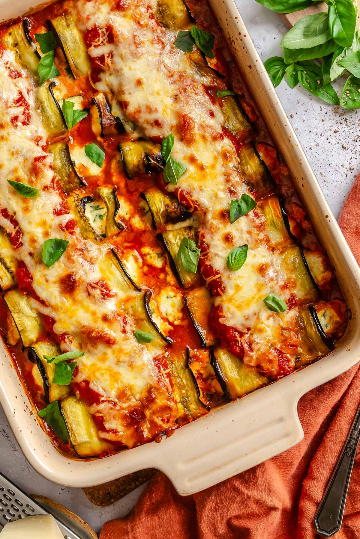 Overhead view of baked eggplant rollatini in a cream baking dish, topped with melted mozzarella, marinara, and fresh basil leaves.