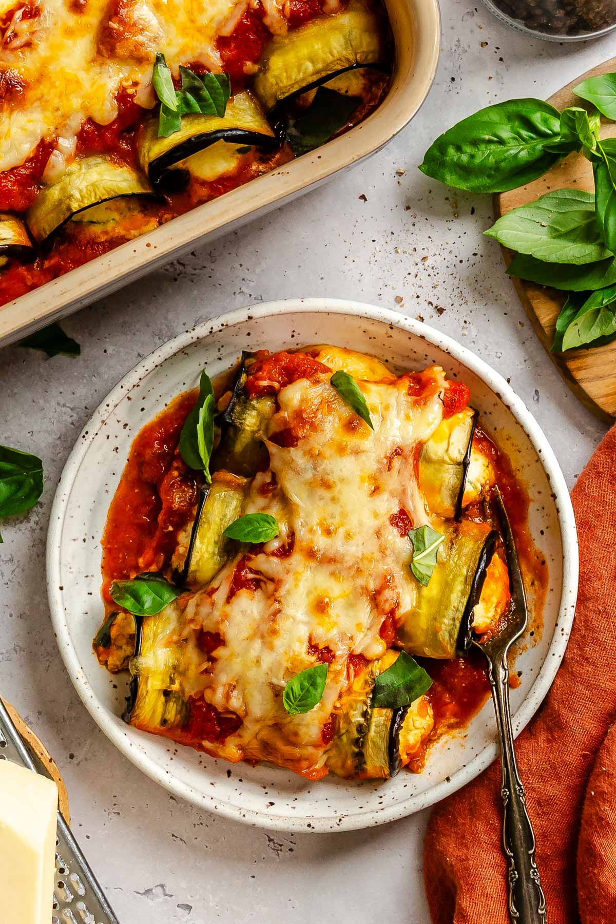 A serving of eggplant rollatini on a speckled plate with marinara and basil, baking dish in the background.