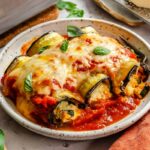 Close-up of a plate of eggplant rollatini with creamy ricotta filling spilling out, topped with marinara and basil.