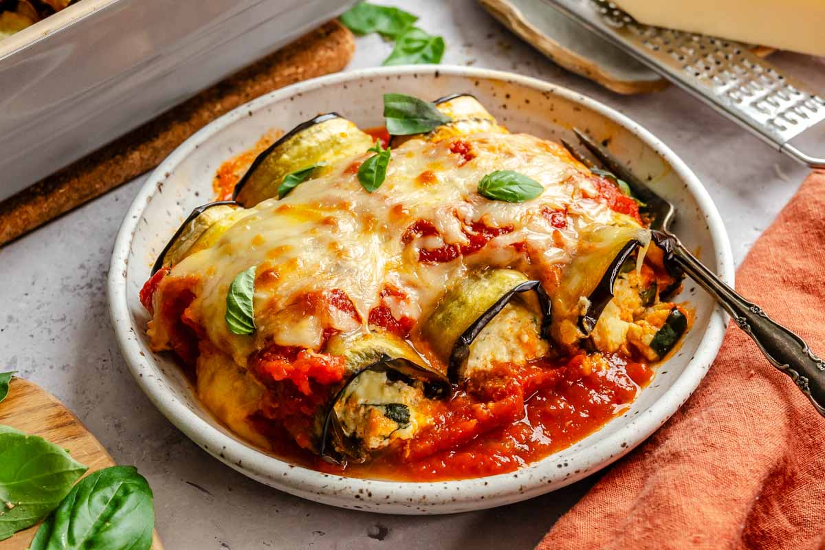 Close-up of a plate of eggplant rollatini with creamy ricotta filling spilling out, topped with marinara and basil.