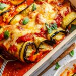 Close-up of eggplant rollatini in a baking dish, melted cheese and marinara glistening, with a serving spoon lifting a piece.