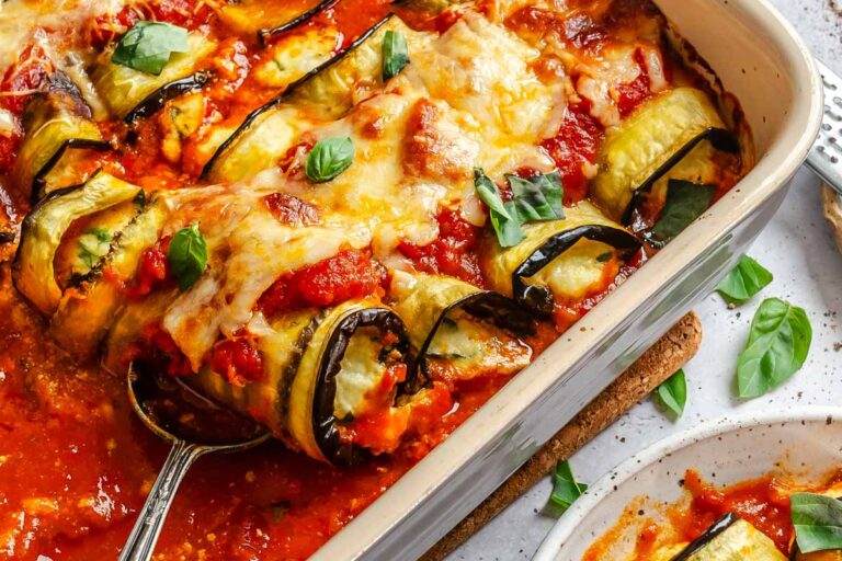 Close-up of eggplant rollatini in a baking dish, melted cheese and marinara glistening, with a serving spoon lifting a piece.