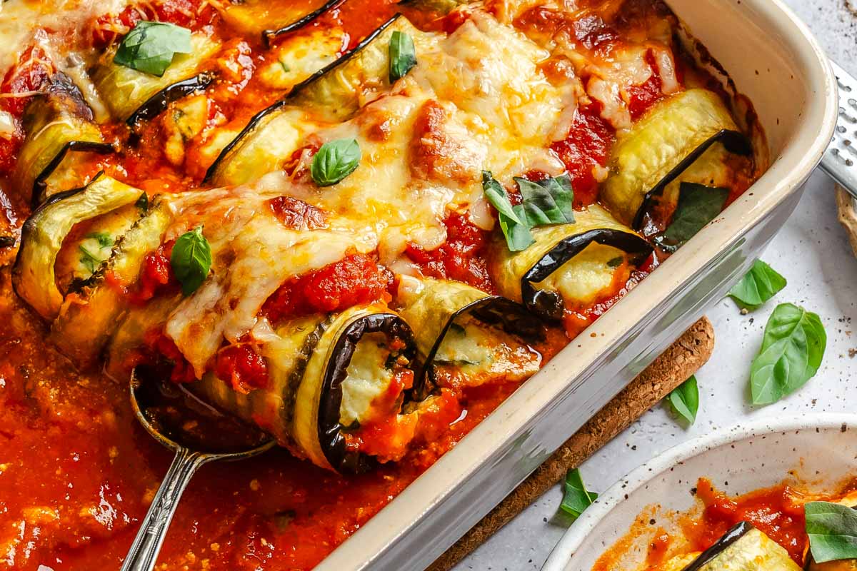 Close-up of eggplant rollatini in a baking dish, melted cheese and marinara glistening, with a serving spoon lifting a piece.