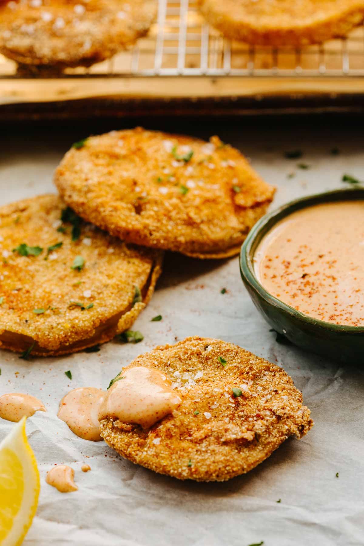 Close-up of stacked fried green tomato slices garnished with herbs, one slice topped with dipping sauce, next to a bowl of sauce and lemon wedges.