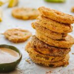 Tall stack of fried green tomato slices on parchment paper with a bowl of dipping sauce, spice blend, and lemon wedges in the background.