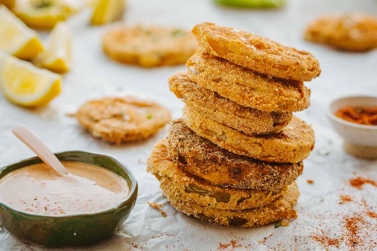 Tall stack of fried green tomato slices on parchment paper with a bowl of dipping sauce, spice blend, and lemon wedges in the background.