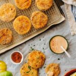 Fried green tomato slices on a wire rack next to a bowl of creamy dipping sauce, a small dish of spice blend, fresh green tomatoes, and lemon wedges arranged on parchment paper.