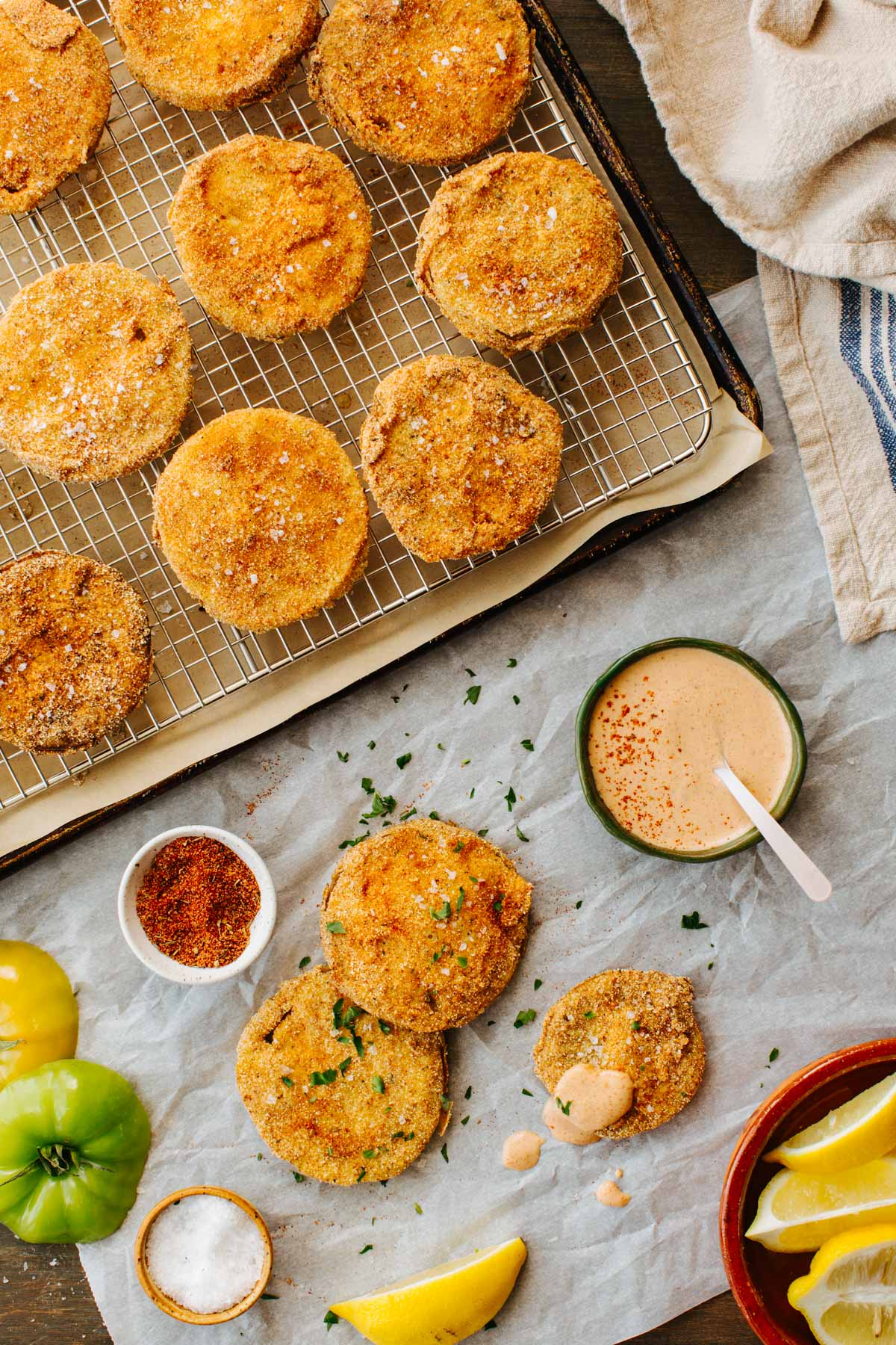 Fried green tomato slices on a wire rack next to a bowl of creamy dipping sauce, a small dish of spice blend, fresh green tomatoes, and lemon wedges arranged on parchment paper. 