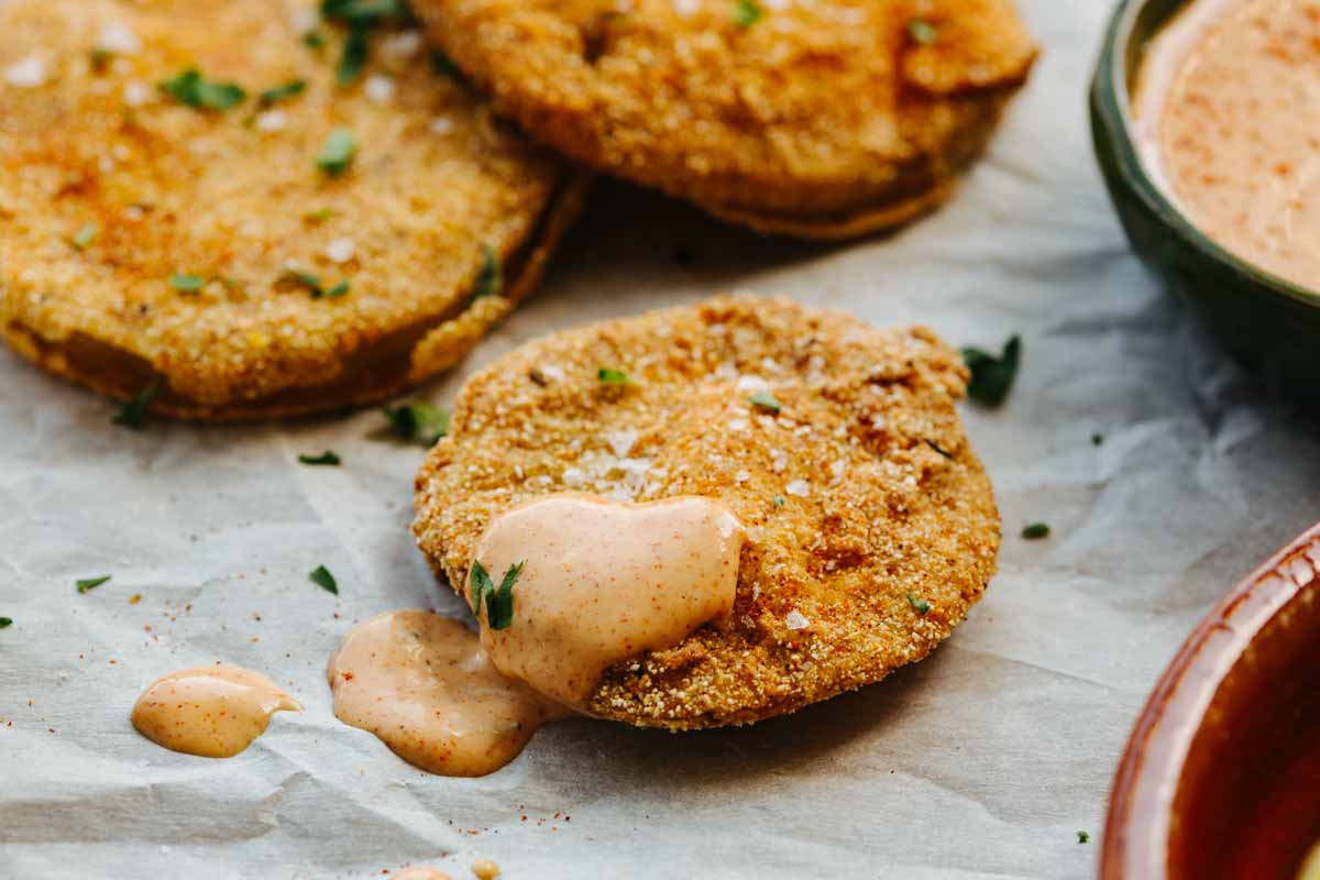 Close-up of a fried green tomato slice topped with a dollop of creamy pink dipping sauce and chopped herbs, with more slices and sauce blurred in the background.