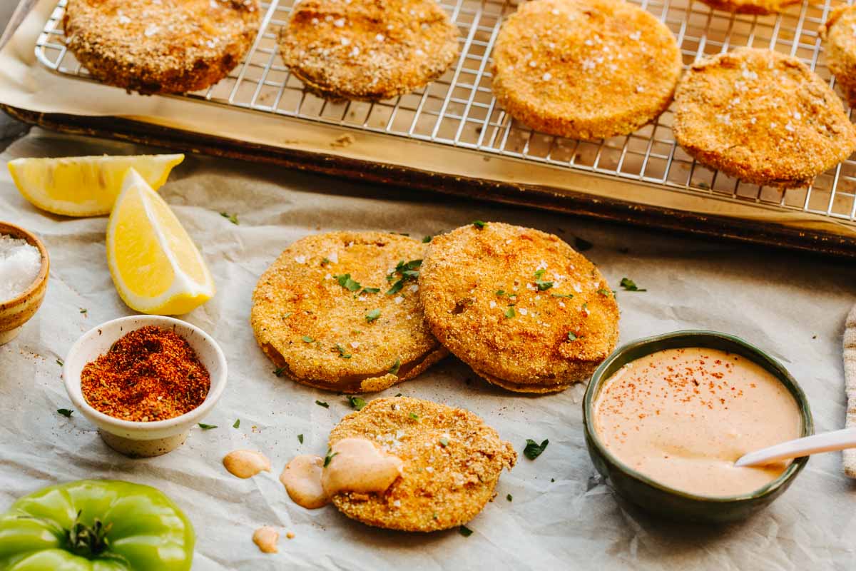 Fried green tomato slices garnished with herbs and flaky salt arranged on parchment paper with lemon wedges, spice blend, and a bowl of dipping sauce, wire rack of tomatoes in the background.