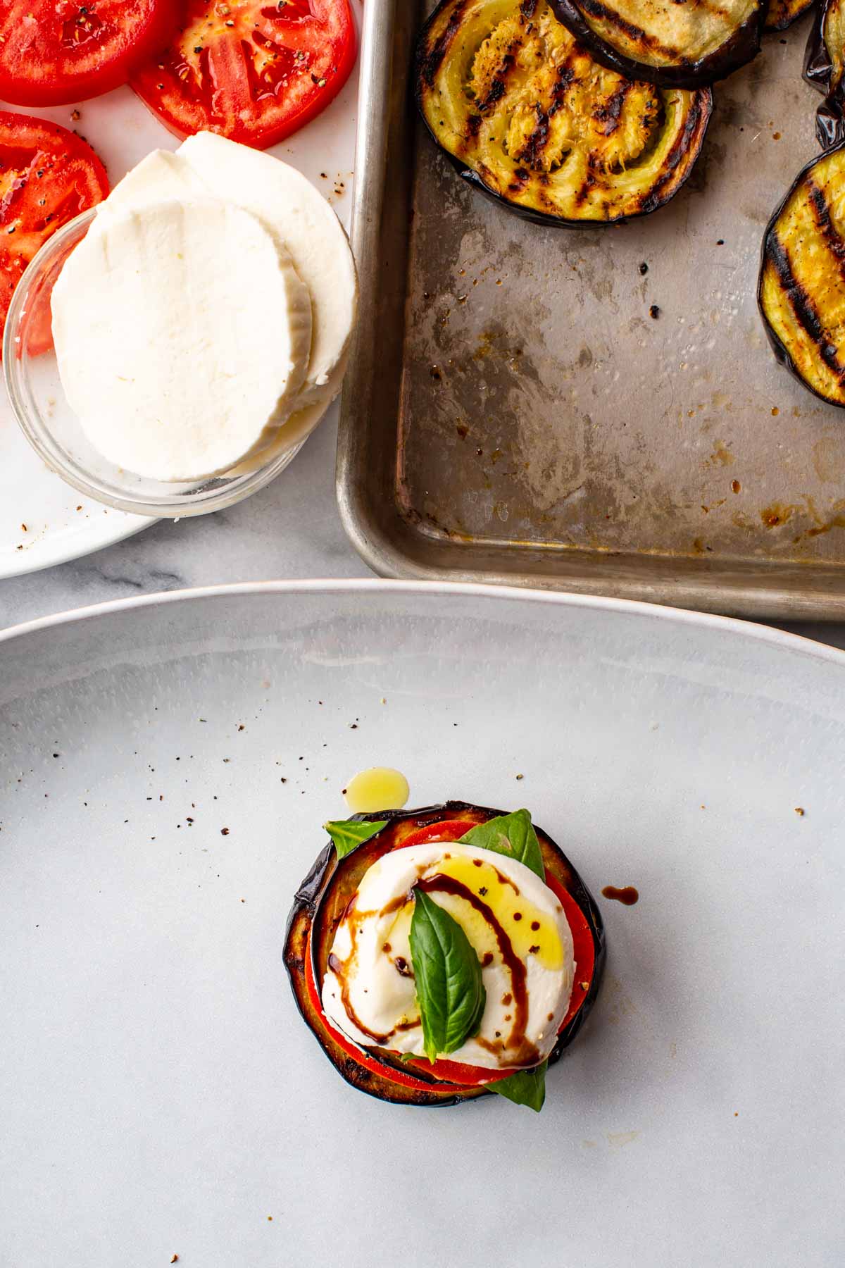 Grilled eggplant caprese stack with tomato, mozzarella, and basil on a plate, with grilled eggplant slices and mozzarella in the background.