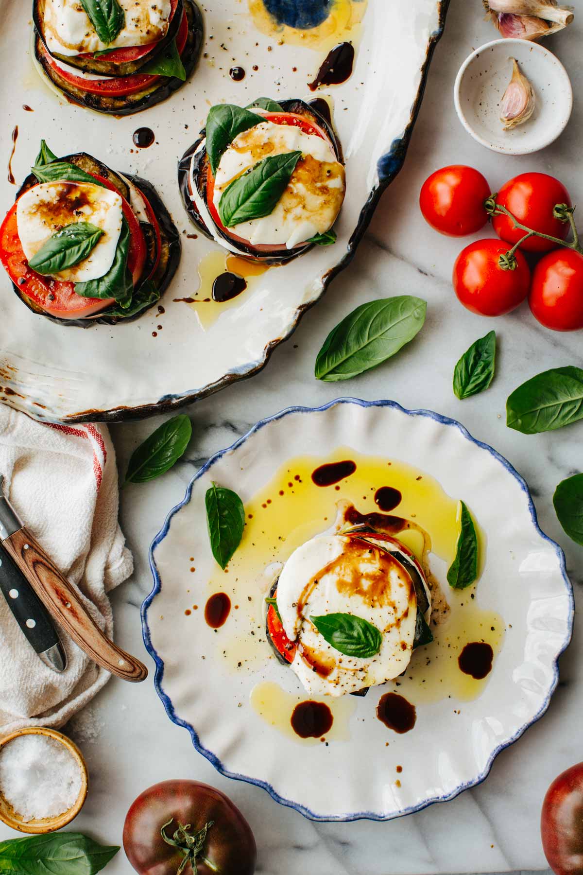 Three plated eggplant caprese stacks drizzled with balsamic, surrounded by fresh tomatoes, basil, and garlic on a marble counter.