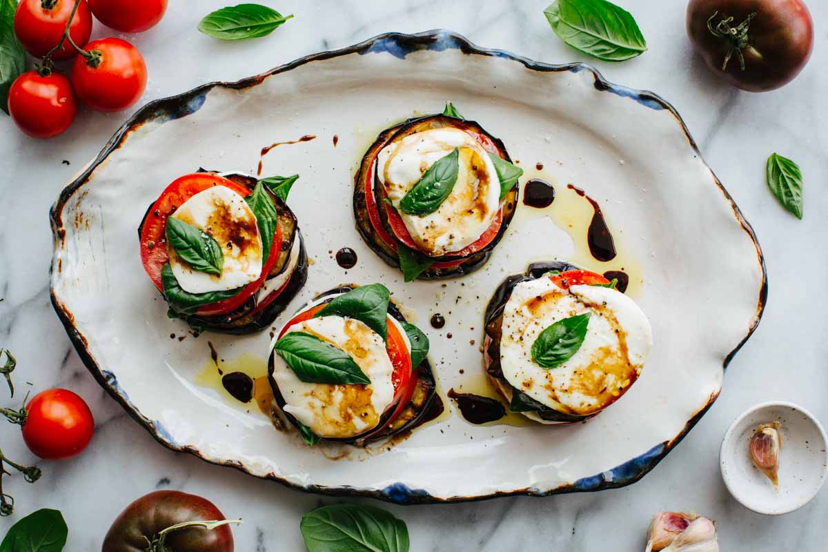 Overhead shot of four eggplant caprese stacks arranged on an oval platter, garnished with basil and balsamic drizzle.