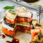 Eggplant caprese stack with a wedge cut out, revealing the layered interior, next to a fork on a decorative plate.