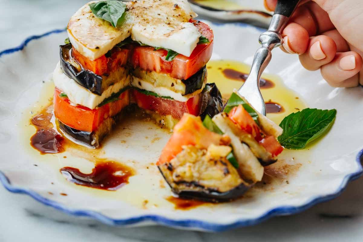 Hand using a fork to take a bite from an eggplant caprese stack with a slice removed on the plate.