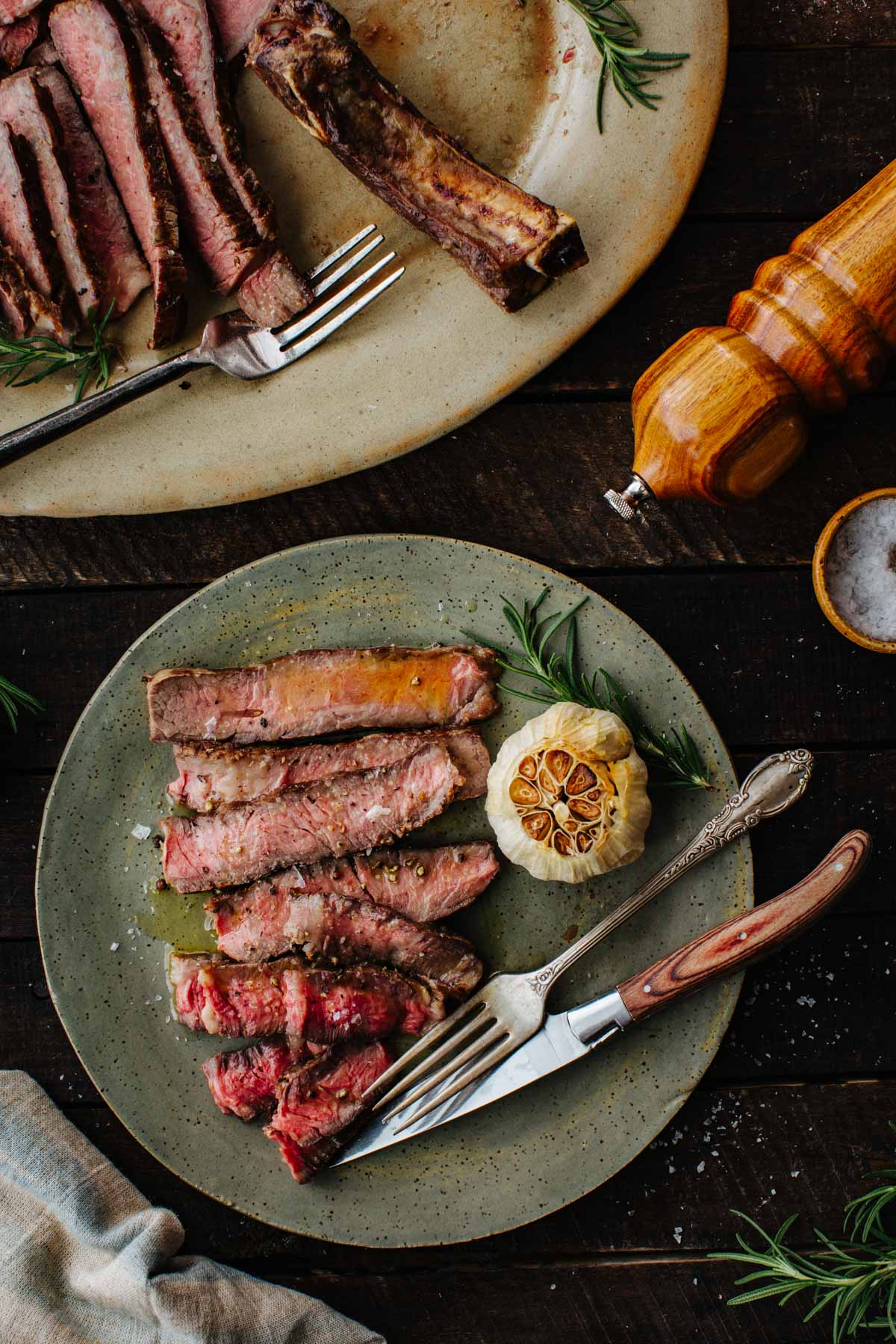 Sliced ribeye steak on a small plate beside a halved roasted garlic bulb and rosemary, with the platter and pepper mill in the background.