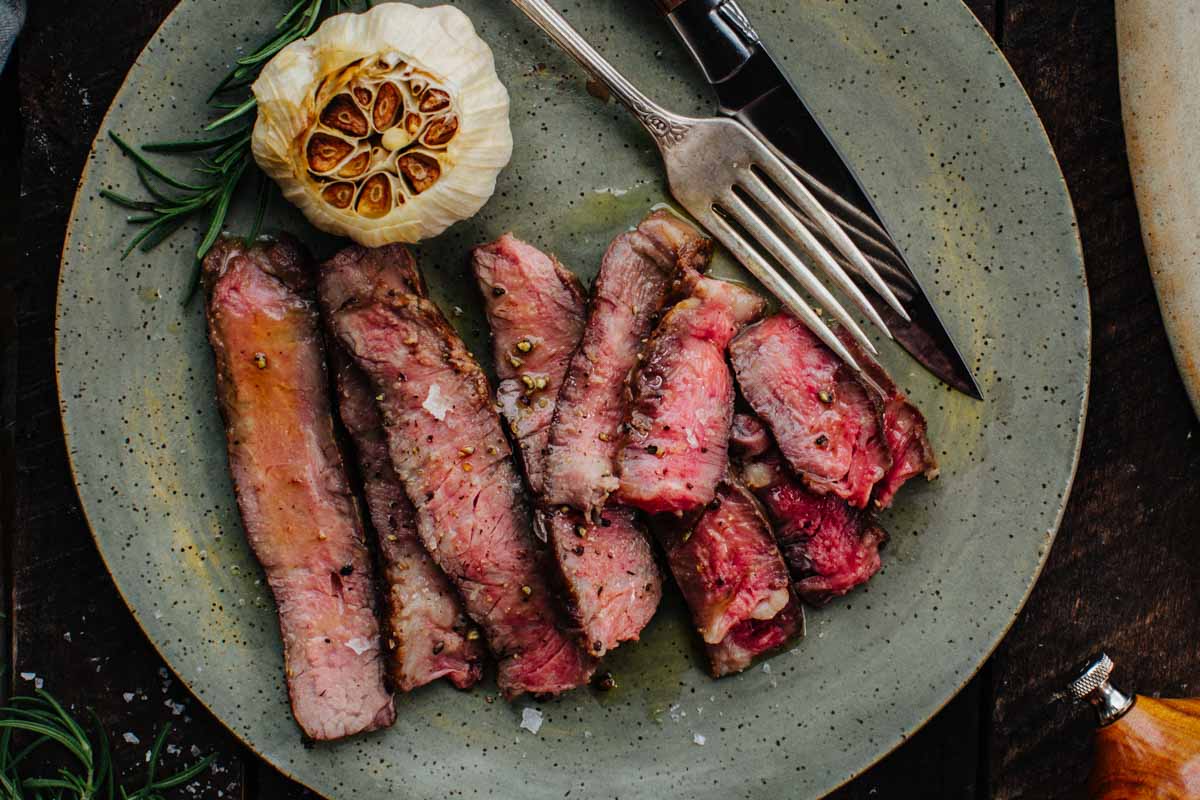 Close-up of sliced medium-rare ribeye steak on a plate with roasted garlic, rosemary, and a fork.