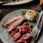 Overhead close-up of sliced ribeye steak with a fork and knife, roasted garlic, and rosemary on a speckled plate.