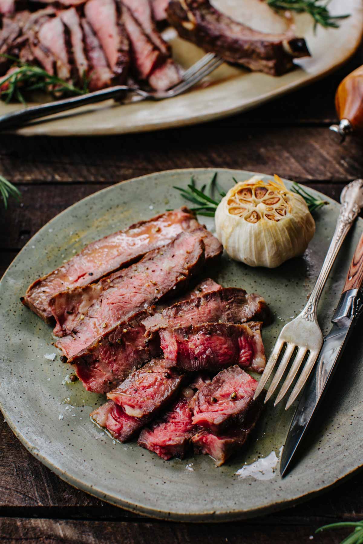 Overhead close-up of sliced ribeye steak with a fork and knife, roasted garlic, and rosemary on a speckled plate.