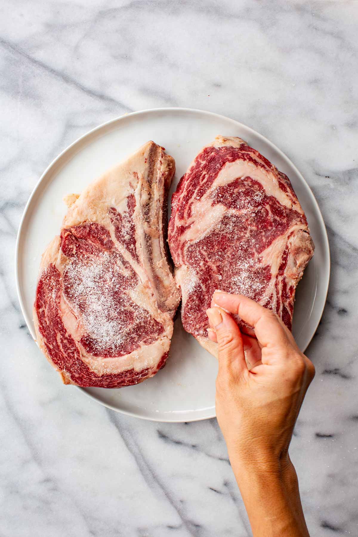 Two raw bone-in ribeye steaks on a white plate, seasoned with salt, with a hand pressing salt into one steak.