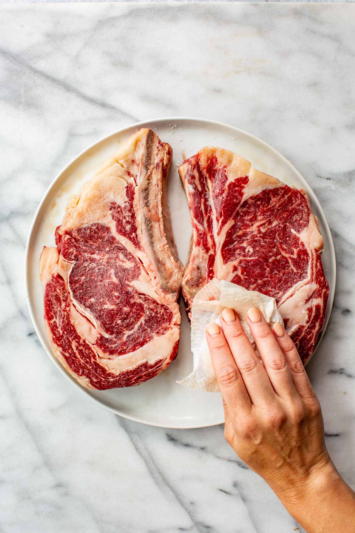 Raw bone-in ribeye steaks on a white plate, being patted dry with a paper towel before grilling.