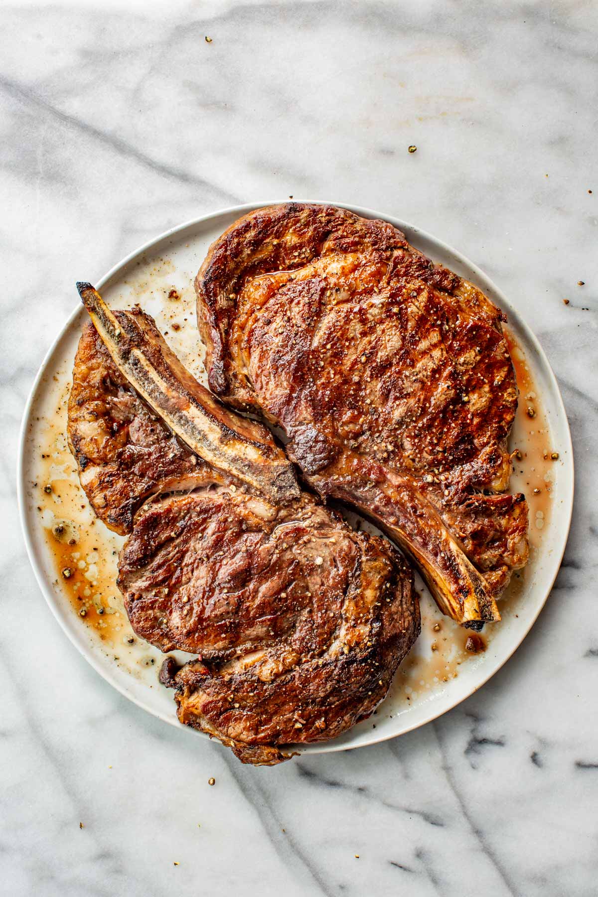 Two grilled bone-in ribeye steaks with char marks on a white plate, bones crossed in the center.