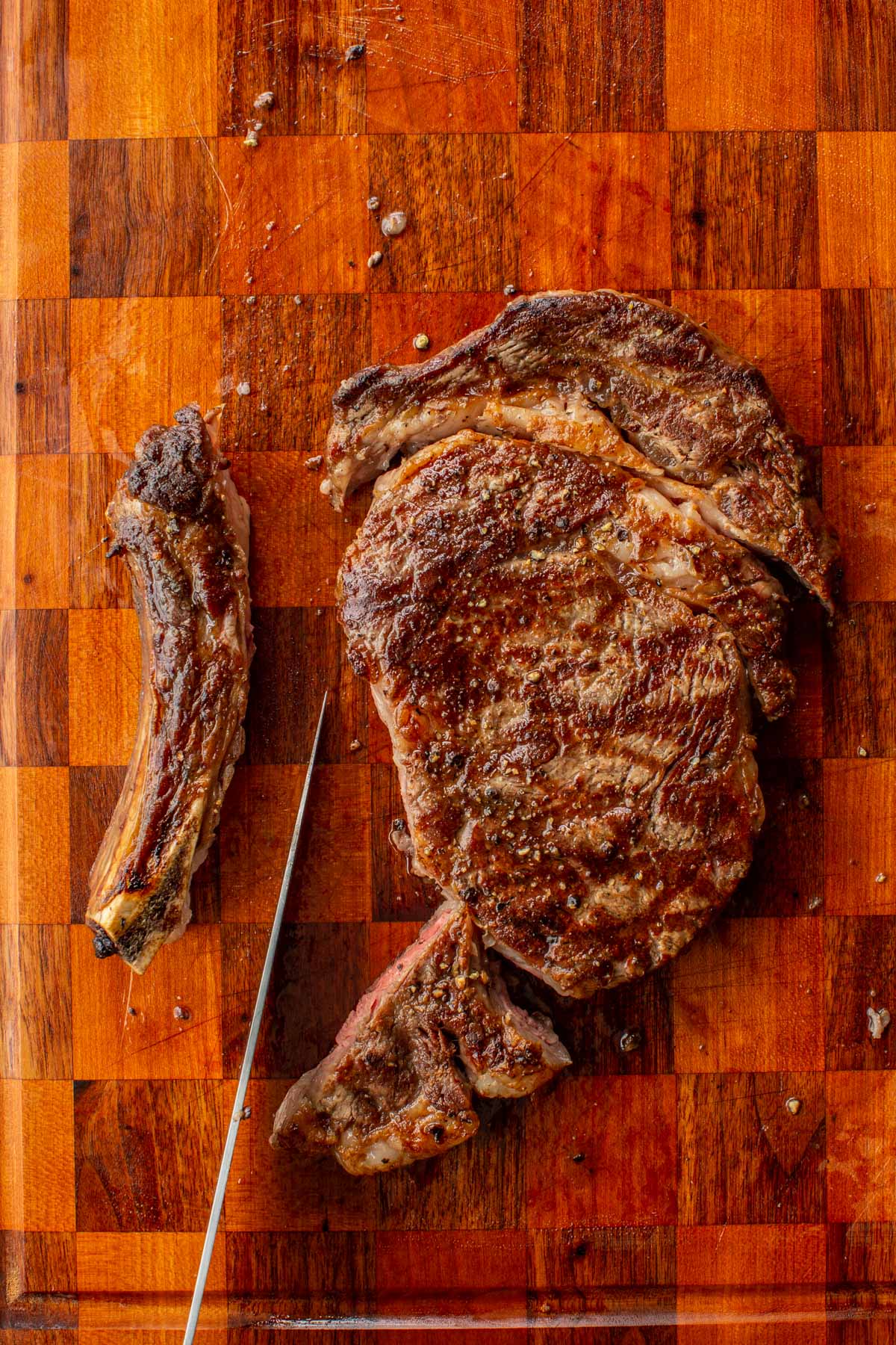 Grilled ribeye steak and bone on a wooden cutting board, with a knife slicing off the first piece.