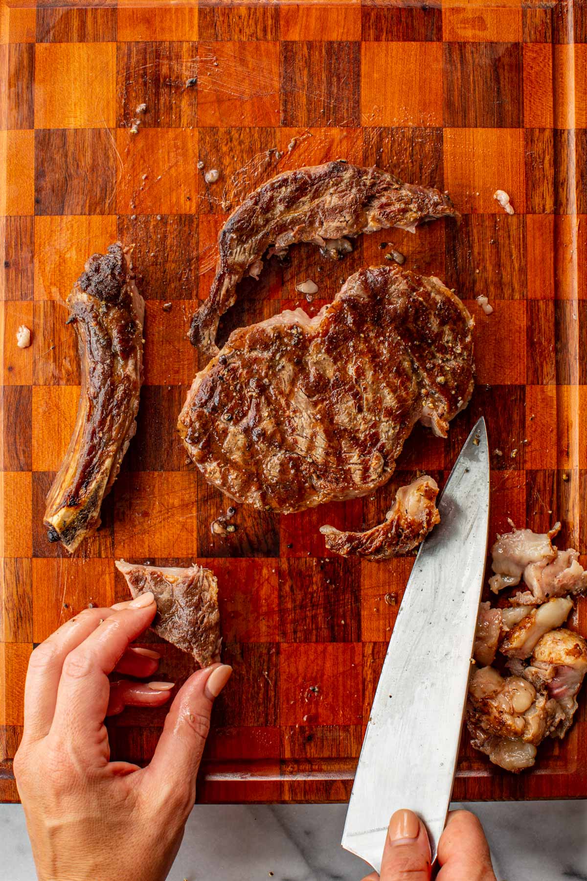 Grilled ribeye steak on a cutting board with the bone removed and fat trimmed to the side, a hand holding a trimmed piece.