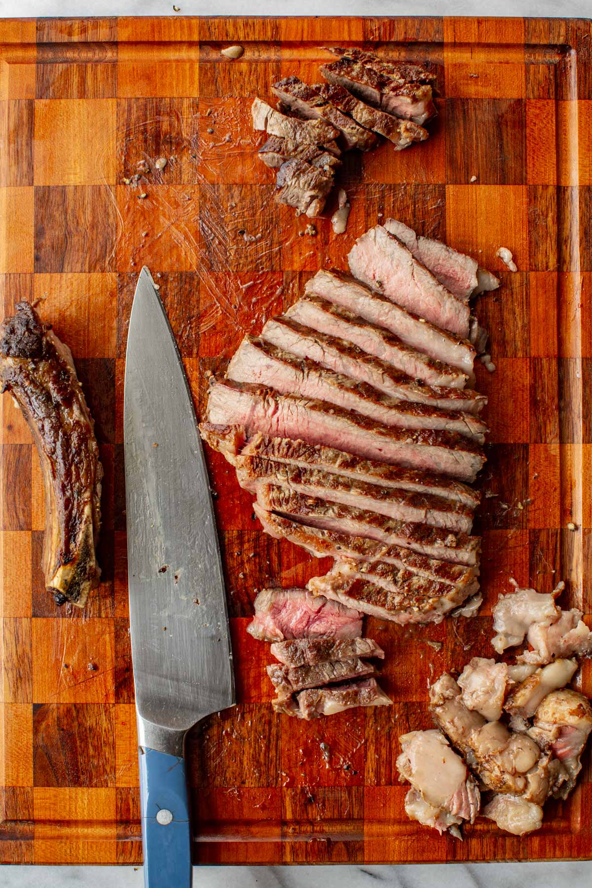 Grilled ribeye steak sliced into strips on a wooden cutting board, showing a pink medium-rare center, with bone and trimmed fat set aside.