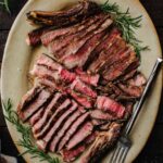 Sliced grilled ribeye steak fanned out on an oval platter with fresh rosemary sprigs, flaky salt, and a fork.