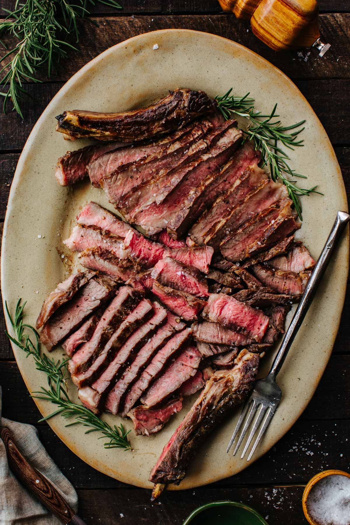 Sliced grilled ribeye steak fanned out on an oval platter with fresh rosemary sprigs, flaky salt, and a fork.