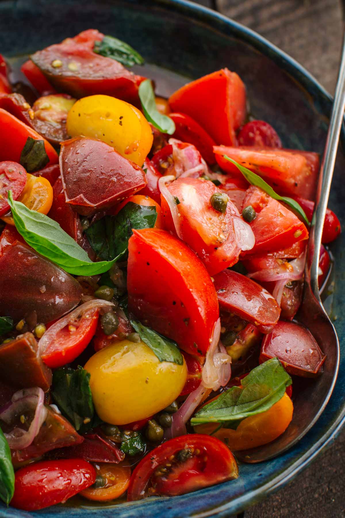 Close-up of a blue ceramic bowl filled with marinated tomato salad and a spoon resting on the plate.