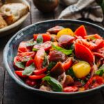 Side view of Italian tomato salad served in a blue ceramic bowl with a spoon in a dark wooden background with bread on the side.