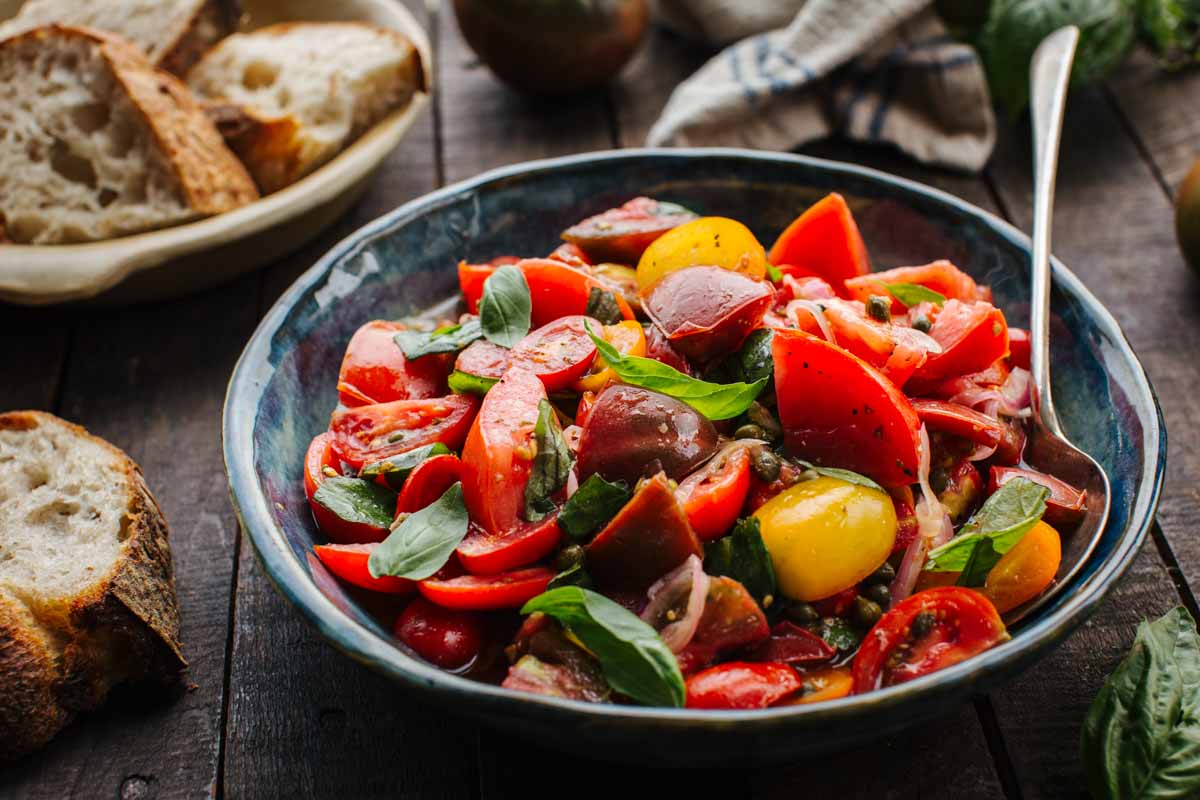 Side view of Italian tomato salad served in a blue ceramic bowl with a spoon in a dark wooden background with bread on the side.