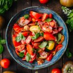 A blue ceramic bowl filled with marinated tomato salad in a dark wooden background surrounded by fresh basil and tomatoes.
