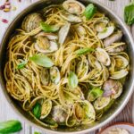 Overhead view of pistachio pesto spaghetti with clams in a skillet, garnished with fresh basil leaves, surrounded by scattered pistachios, basil, and a plate of shaved Parmigiano on a wooden table.