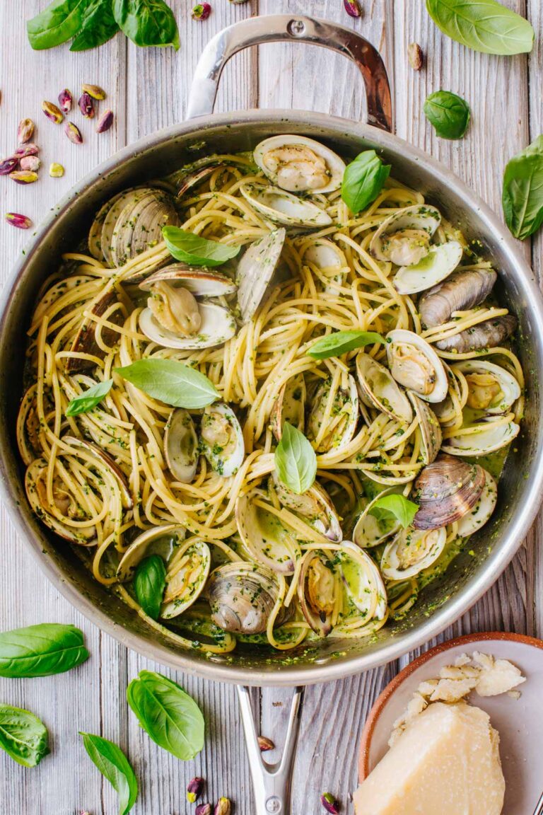Overhead view of pistachio pesto spaghetti with clams in a skillet, garnished with fresh basil leaves, surrounded by scattered pistachios, basil, and a plate of shaved Parmigiano on a wooden table.