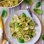 A plated serving of spaghetti with pistachio pesto and clams, garnished with basil leaves and a fork, next to the full skillet, a bowl of pistachios, and a cheese grater on a wooden table.