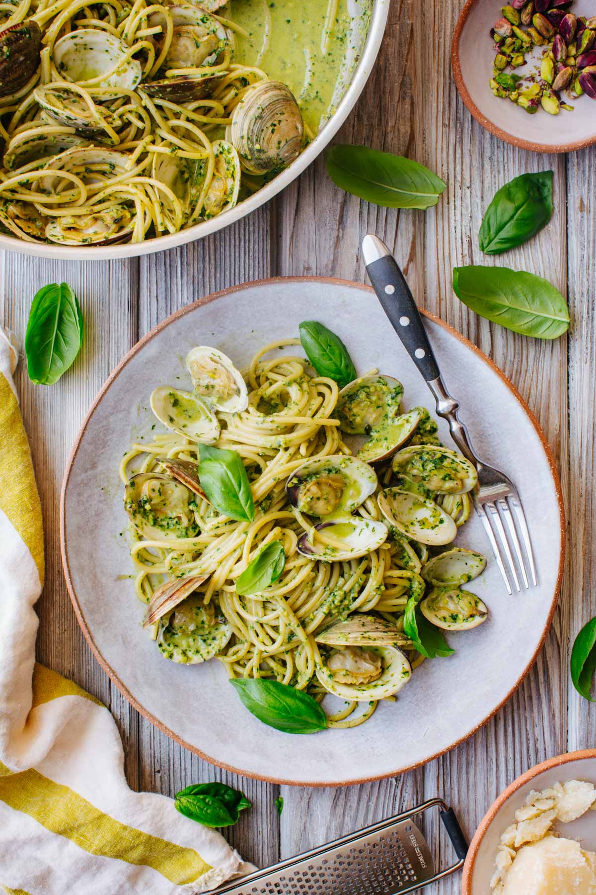 A plated serving of spaghetti with pistachio pesto and clams, garnished with basil leaves and a fork, next to the full skillet, a bowl of pistachios, and a cheese grater on a wooden table.