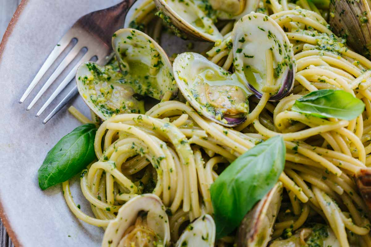 Close-up of a plate of pistachio pesto spaghetti with open clams and basil leaves, with a fork resting on the edge.