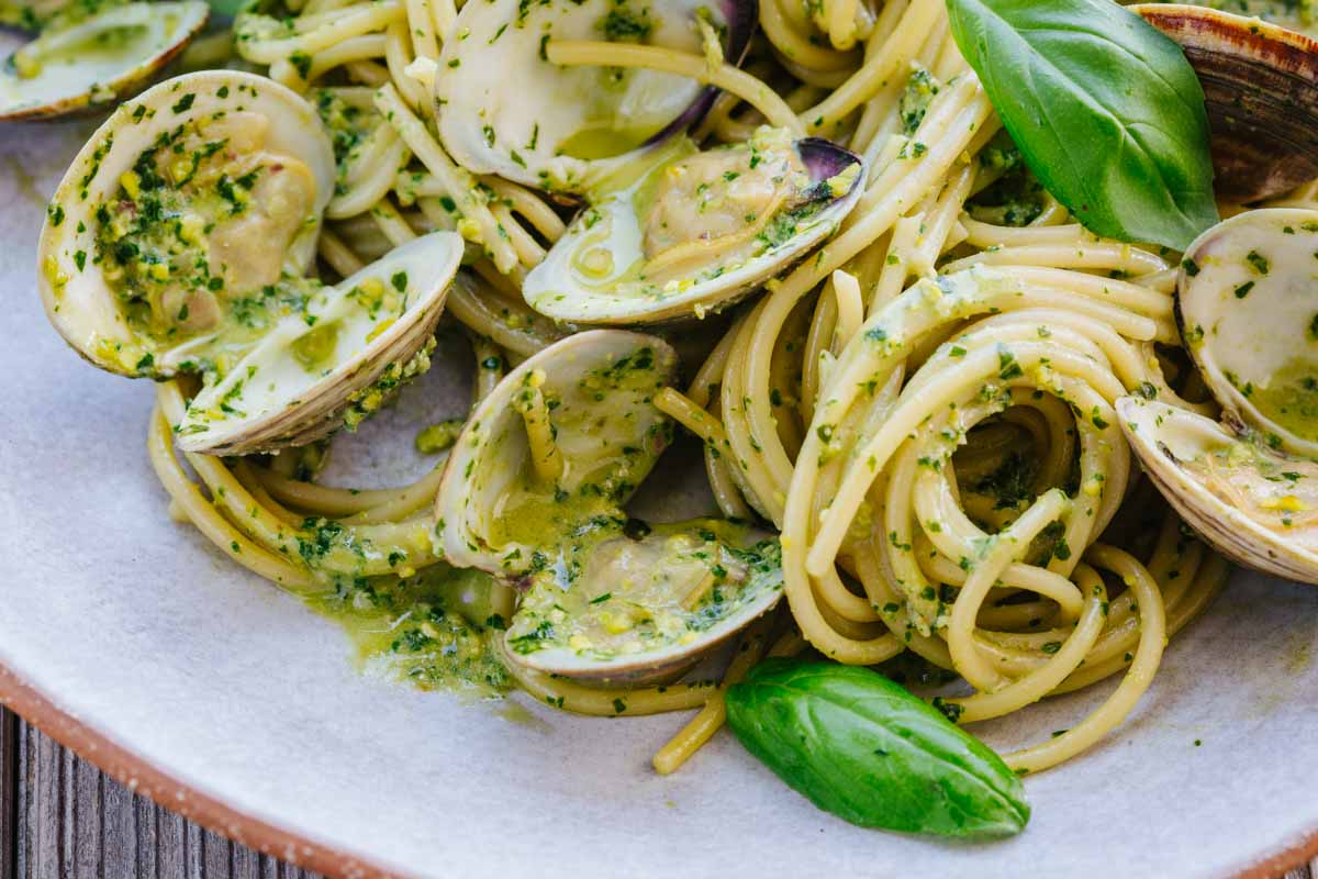 Close-up of pistachio pesto-coated spaghetti with open clams and a basil leaf on a speckled ceramic plate.