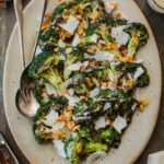 Overhead shot of a full platter of charred grilled broccoli Caesar salad, drizzled with creamy dressing and topped with crunchy croutons and shaved Parmigiano, surrounded by lemon halves, garlic, and small bowls of salt and pepper.
