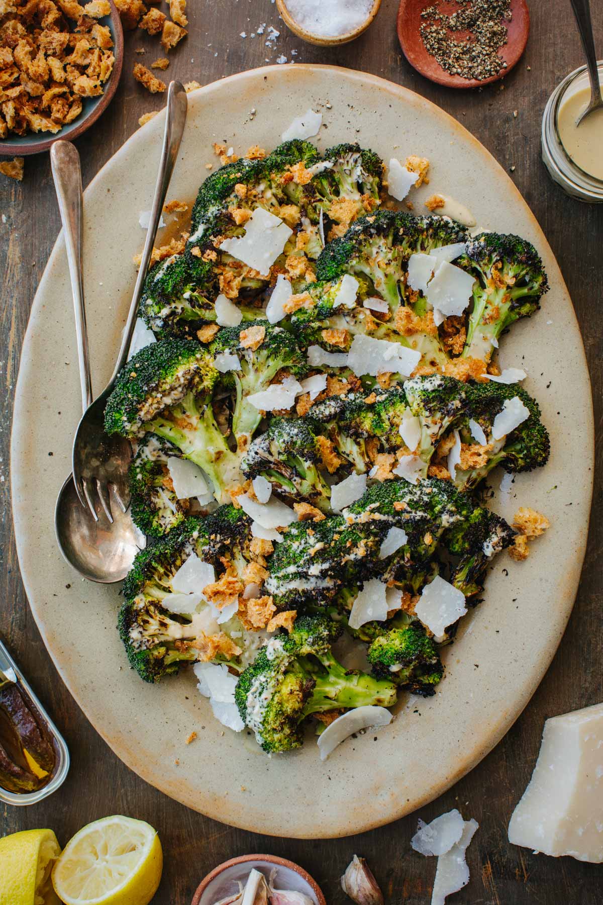 Overhead shot of a full platter of charred grilled broccoli Caesar salad, drizzled with creamy dressing and topped with crunchy croutons and shaved Parmigiano, surrounded by lemon halves, garlic, and small bowls of salt and pepper.