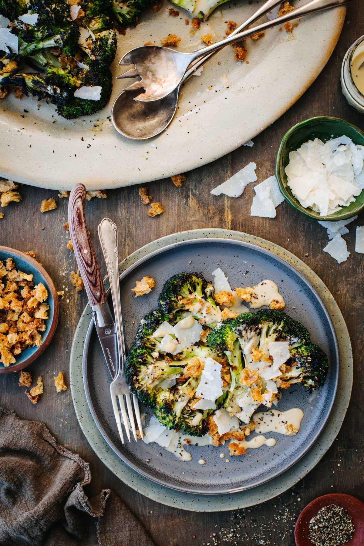 Overhead shot of a single serving of grilled broccoli Caesar salad on a blue plate with a fork and knife, next to the serving platter, a bowl of Parmigiano shavings, and scattered croutons on a wood table.