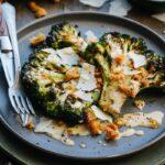 Close-up of a plated portion of charred grilled broccoli drizzled with Caesar dressing and topped with croutons and shaved Parmigiano, fork resting alongside on a dark blue plate.