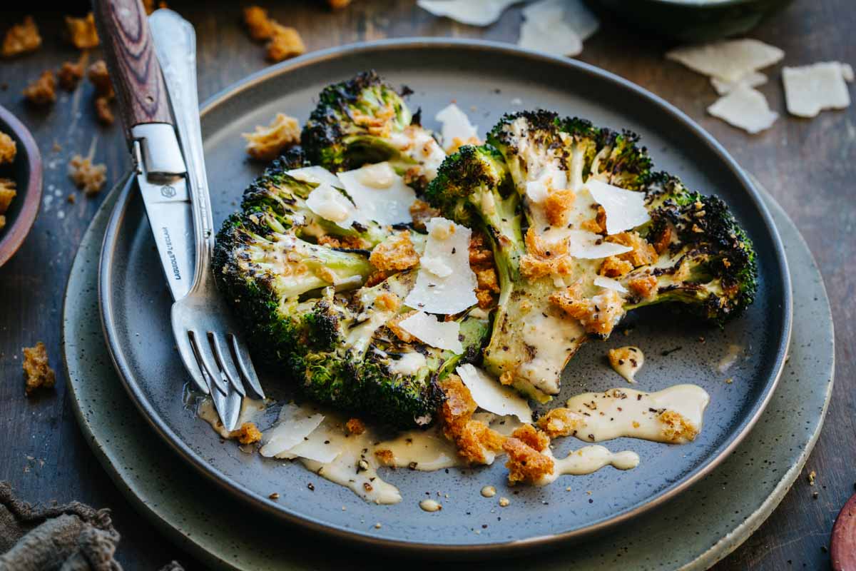Close-up of a plated portion of charred grilled broccoli drizzled with Caesar dressing and topped with croutons and shaved Parmigiano, fork resting alongside on a dark blue plate.